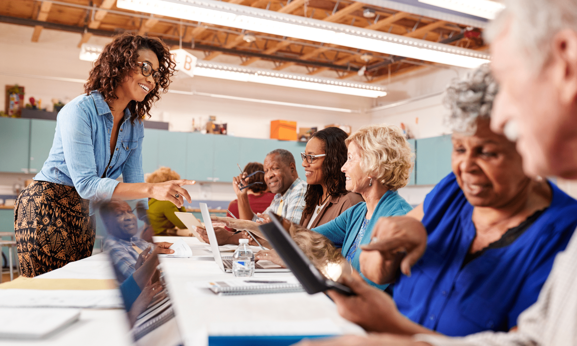A group of older adults seated at a table use digital devices while a woman stands and assists them in a classroom setting.