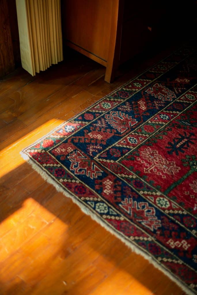 Sunlight falls on a patterned red and blue rug covering a wooden floor next to a wooden cabinet and vertical blinds.