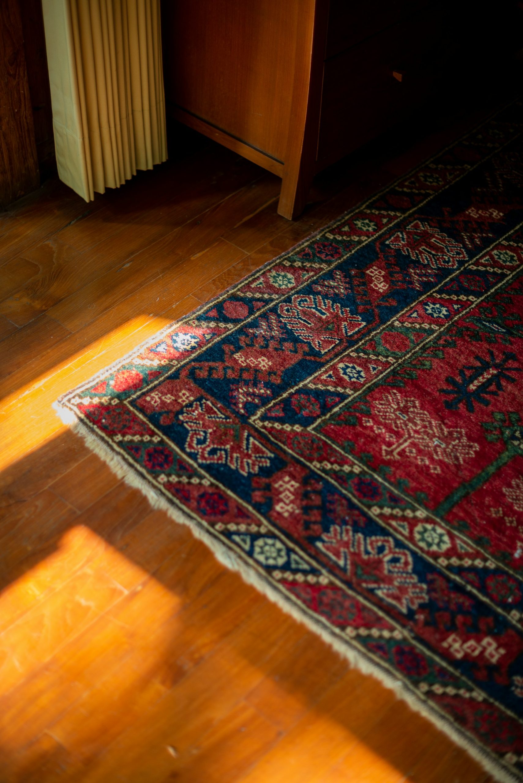 Sunlight falls on a patterned red and blue rug covering a wooden floor next to a wooden cabinet and vertical blinds.